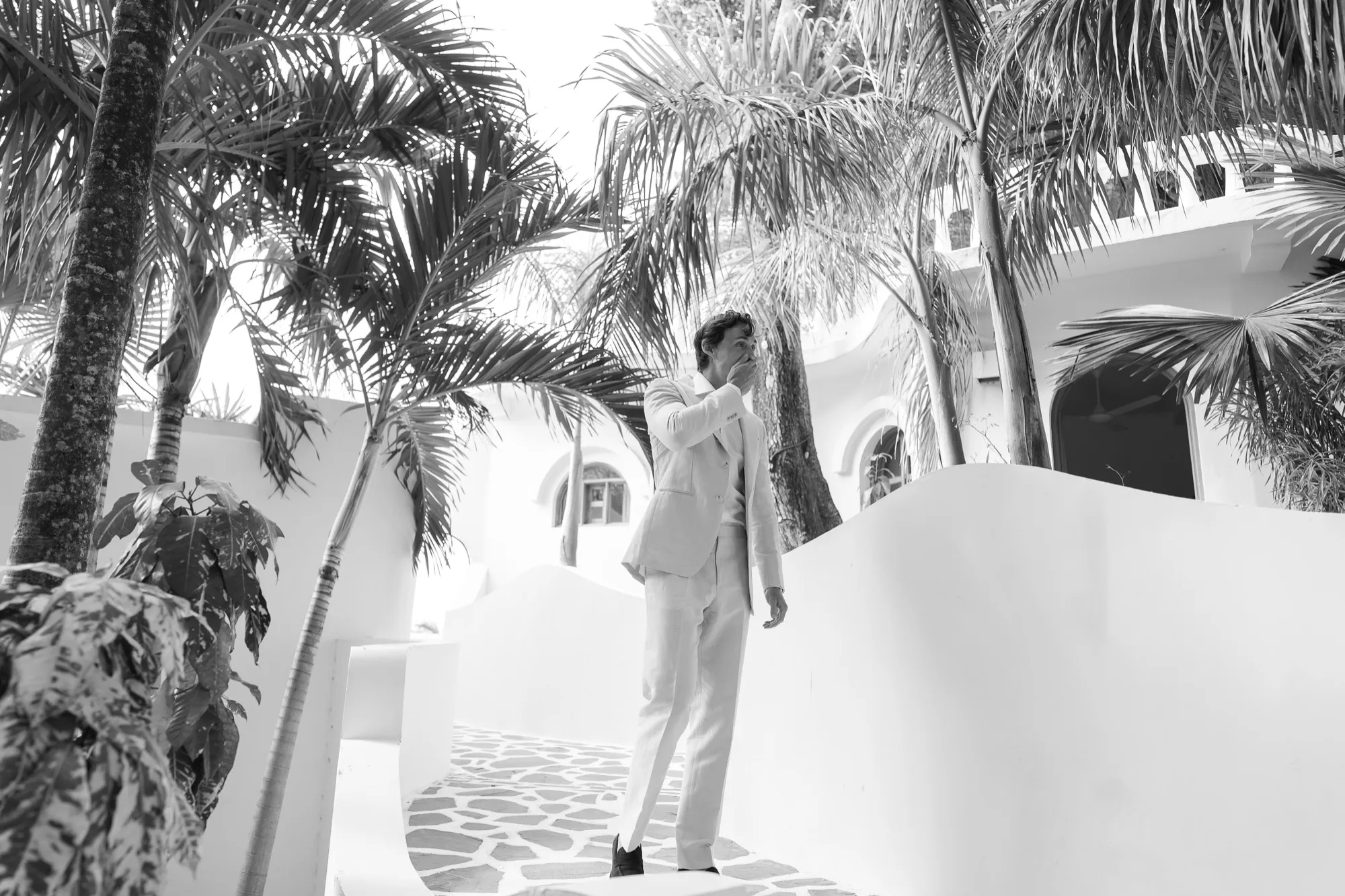 A groom walking among palms beside a white villa, in black and white