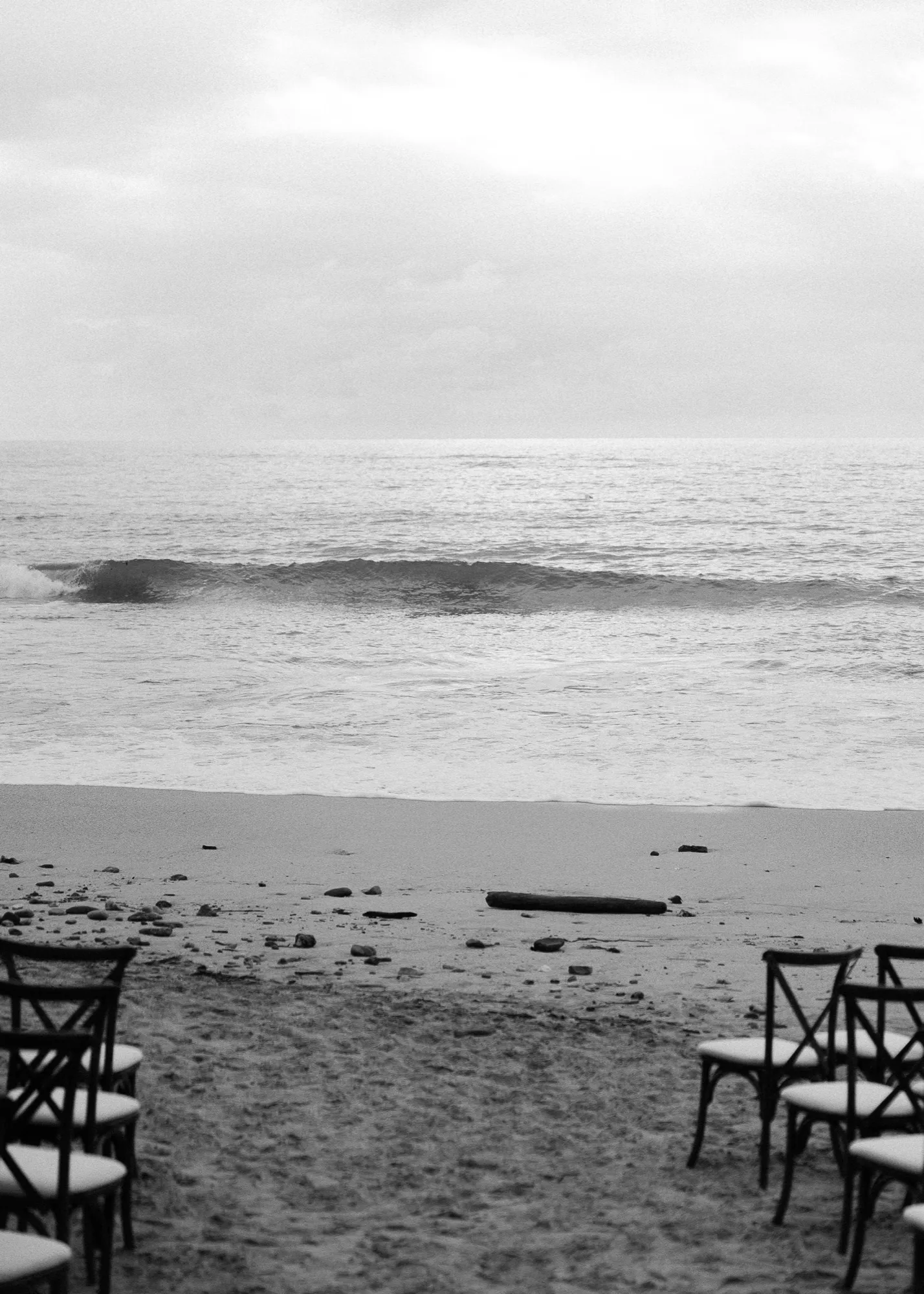 A quiet black-and-white view of a breaking wave beyond ceremony chairs on the beach