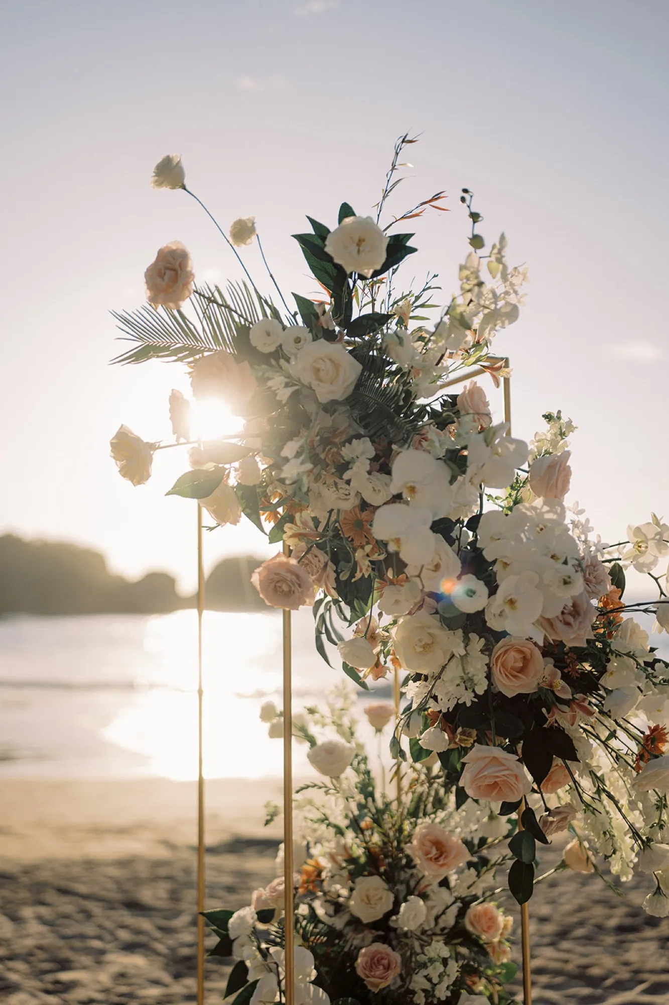 A blush-and-white floral arch backlit by the sunset on the beach