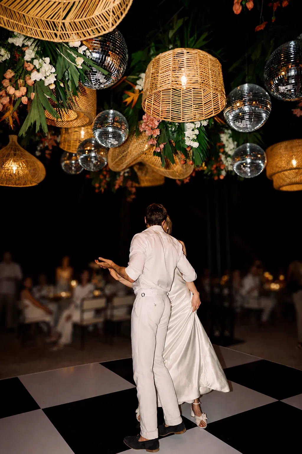 A couple's first dance on a chequerboard floor beneath rattan lanterns, disco balls and tropical florals