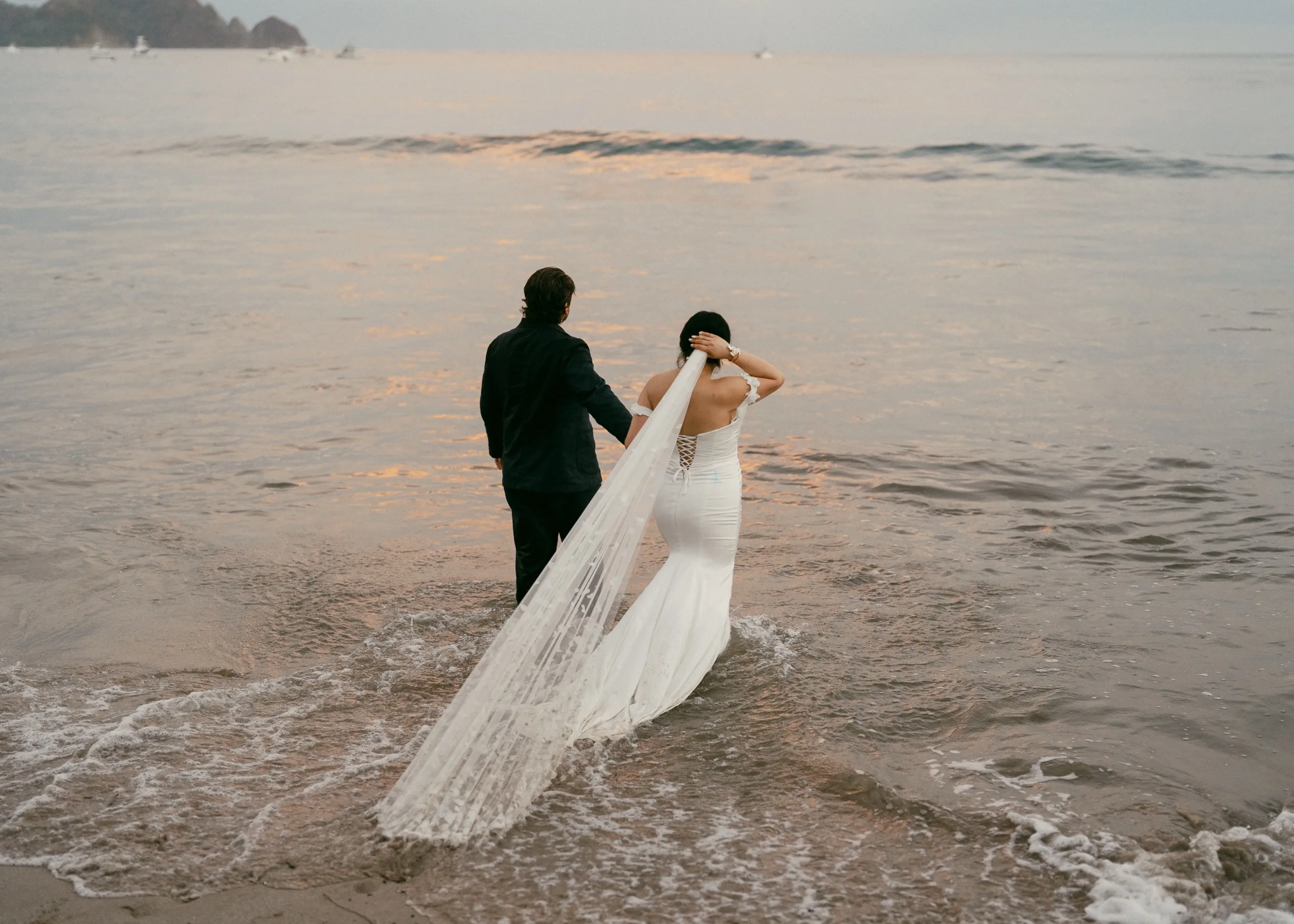 A newly married couple walking into the shallows at dusk on a Nosara beach, the bride's veil trailing behind her