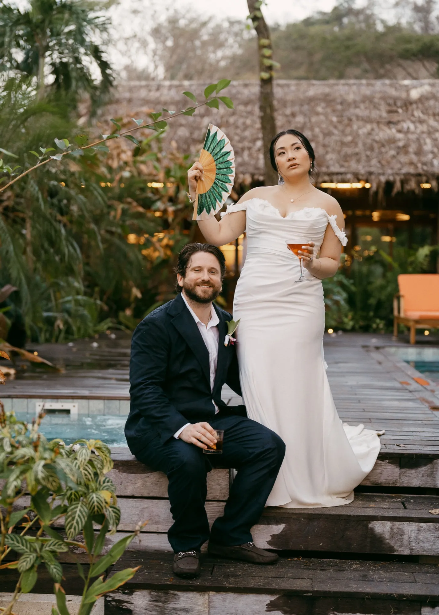 A bride with a hand fan and cocktail beside her seated groom by the pool at a Nosara wedding