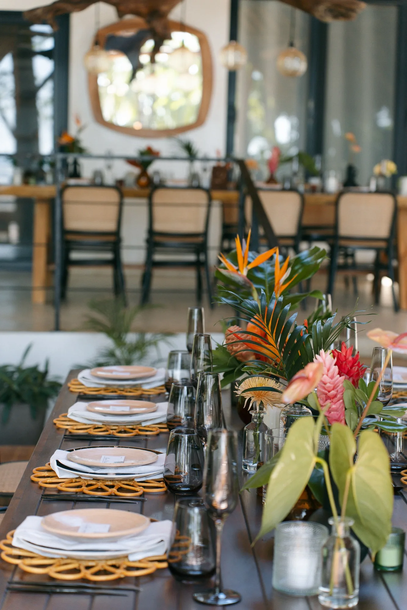 A long reception table set with rattan chargers, smoke-glass stemware, and tropical florals at a Nosara wedding