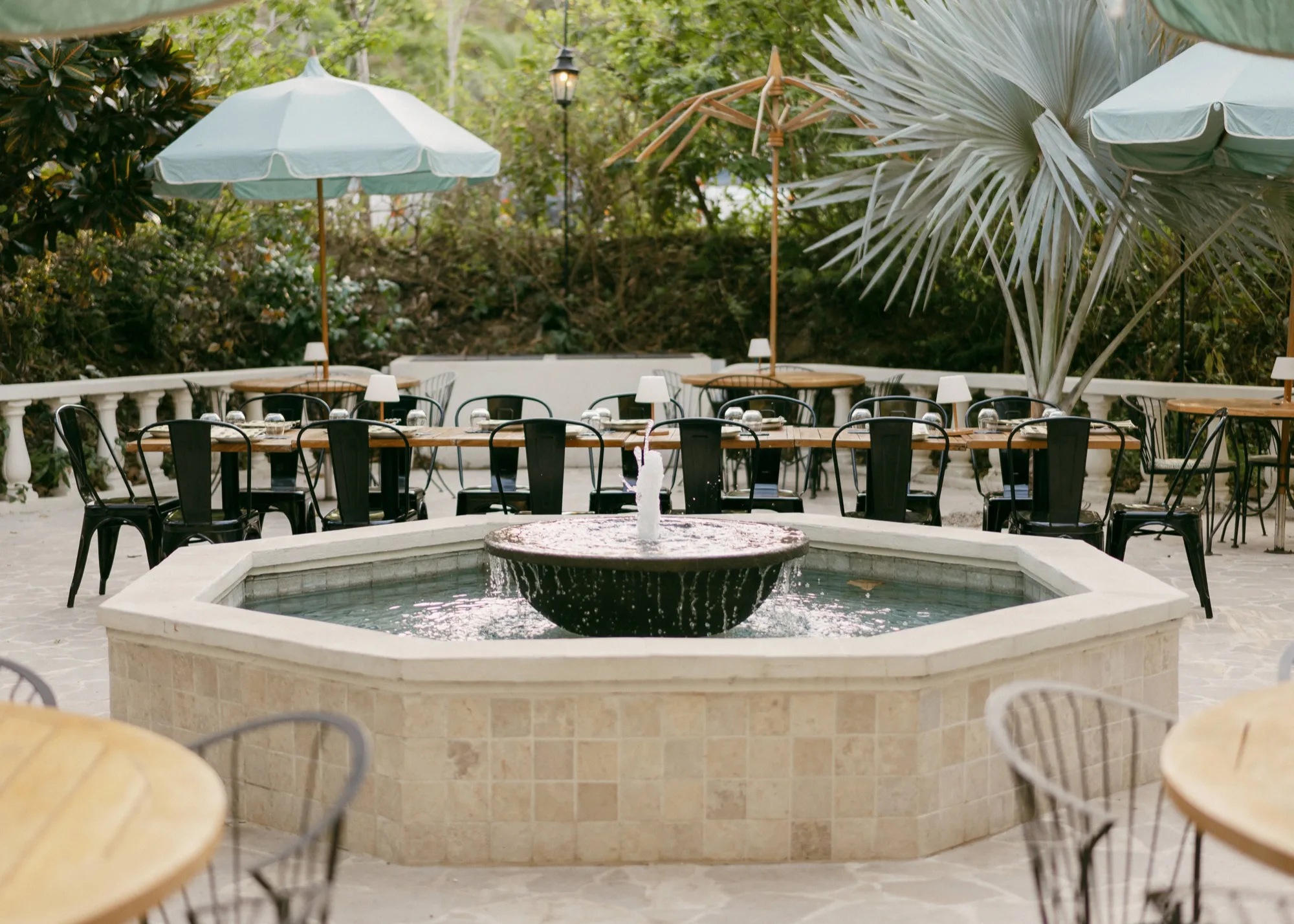A garden courtyard set for a welcome dinner around a stone fountain, shaded by parasols and palms in Nosara